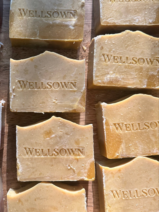 Multiple bars of WELLSOWN handmade soap arranged in vertical columns on a wooden surface. The soaps include marbled purple and cream bars, solid pale yellow bars, and solid golden-yellow bars, each embossed with the brand name.