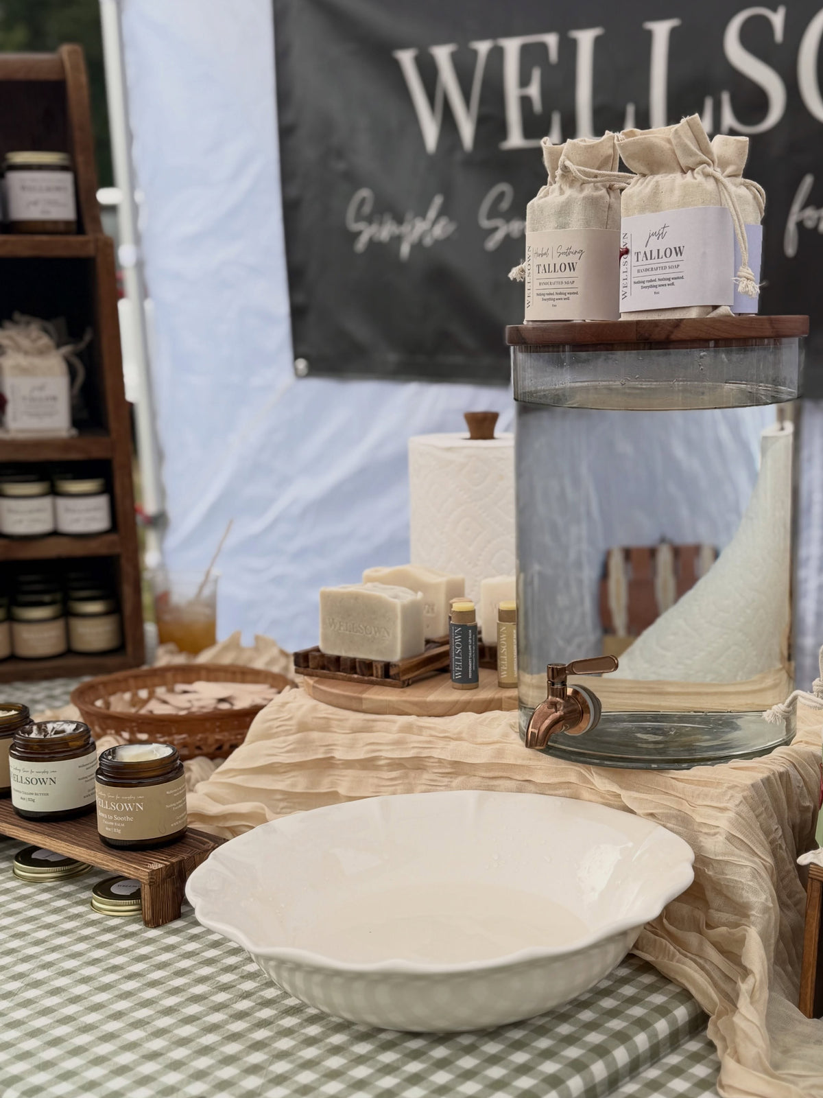 A rustic market stall display featuring handmade soap bars, small glass jars, a large glass water dispenser with a wooden lid, and a white ceramic bowl on a green-and-white checkered tablecloth. A black banner in the background reads 'WELLSOWN.